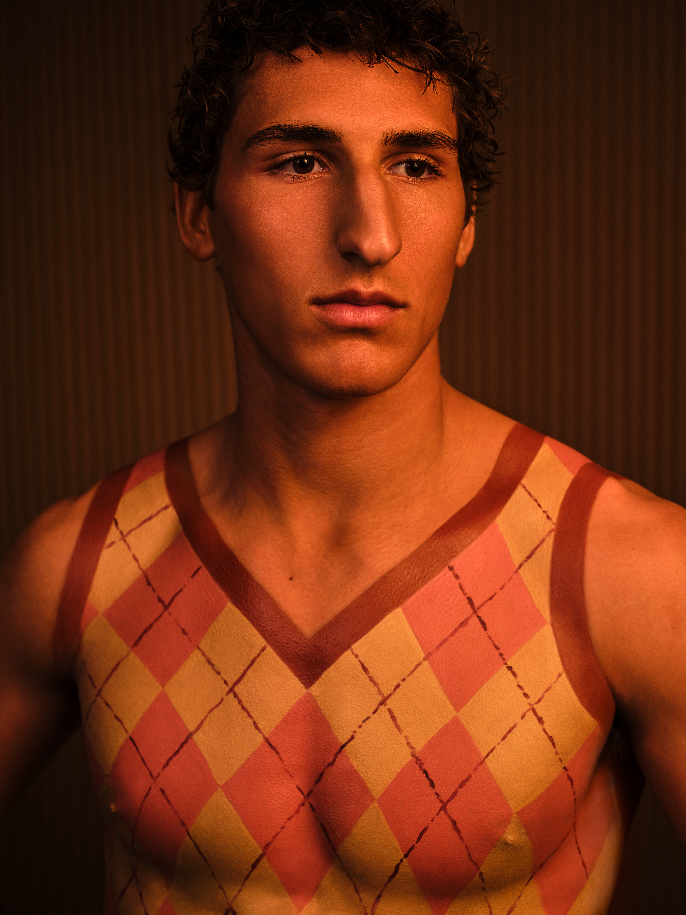 Close up portriat of male model looking past the camera with body a painted plaid sweater top against a dark wood backdrop